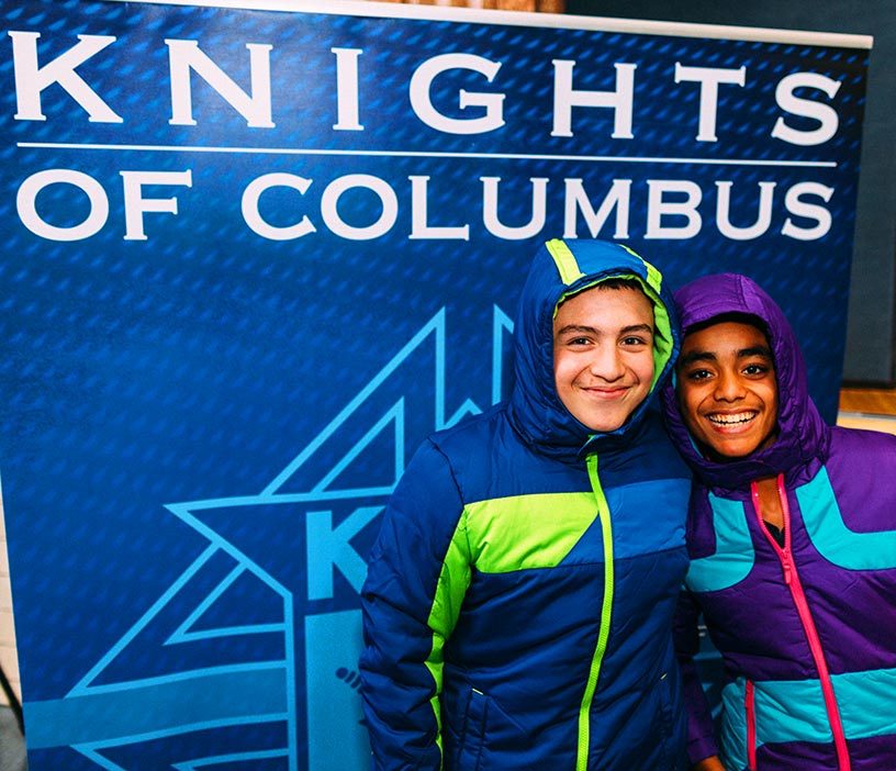 Two smiling children in new winter coats in front of a Knights of Columbus banner