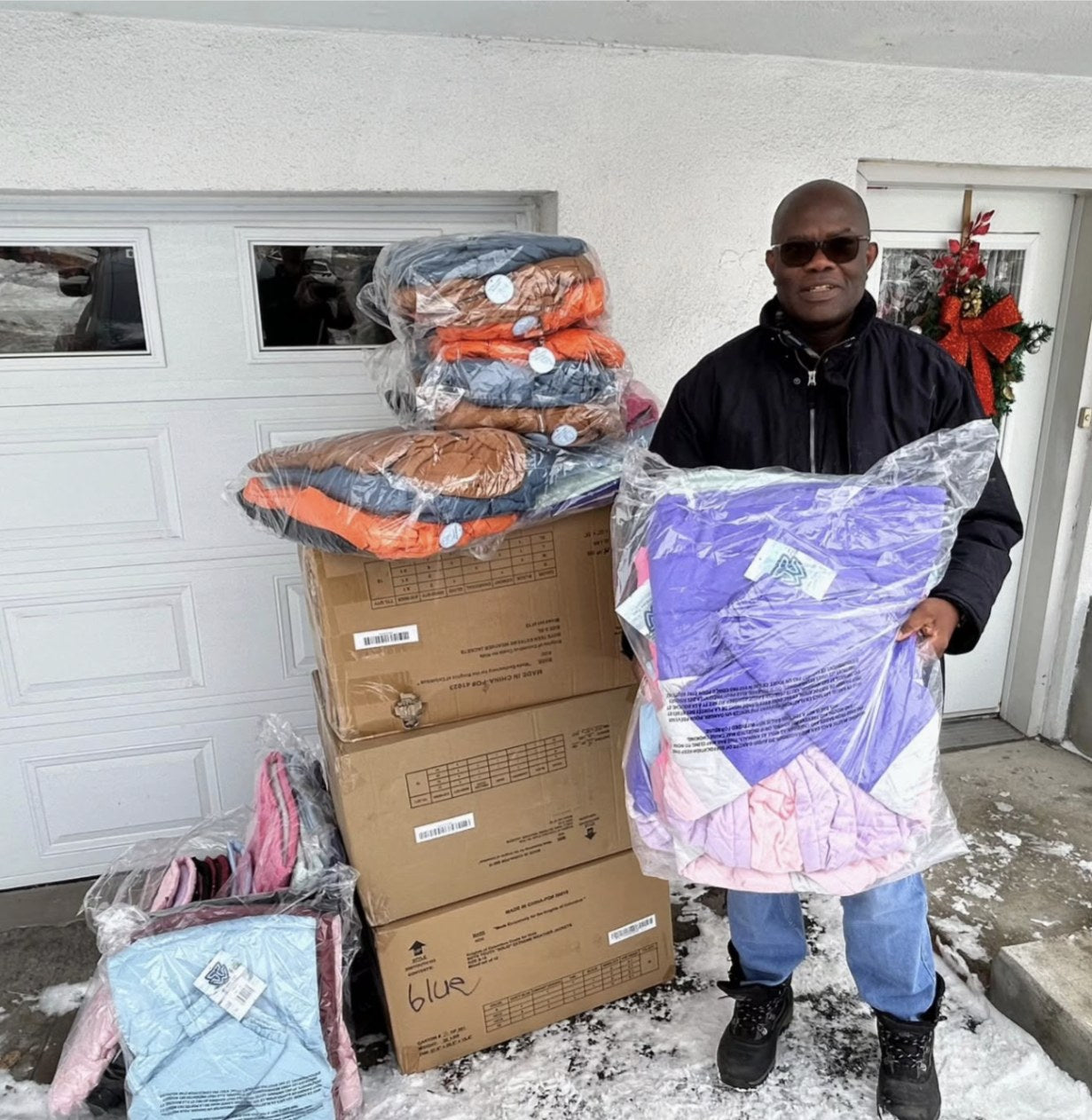 A man holds a bag of new winter coats beside stacked boxes of coats on a snowy doorstep